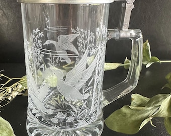 May include: A clear glass beer stein featuring an etched design of ducks in flight over a marshy landscape. The vessel includes a metal lid with a thumb lever and a sturdy handle, with a decorative starburst pattern embossed on the base.