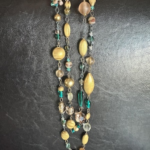 May include: Two strands of beaded necklaces with a variety of beads in gold, clear, and teal. The beads include round, oval, and textured shapes. The necklaces are held up by a hand with pink nail polish.