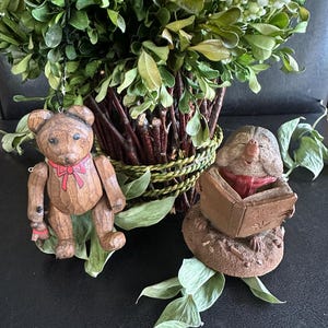May include: A carved wooden teddy bear ornament with a painted red bow and a brown textured figurine of a mole reading a book with a red scarf, both positioned in front of a green leafy plant with white berries and a dark woven basket base.