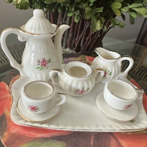 Puede incluir: Un juego de té de porcelana blanca con un diseño floral. El juego incluye una tetera, una jarra de crema, un azucarero, dos tazas de té y dos platillos. El juego está en una bandeja blanca con un borde dorado.