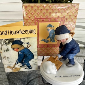 May include: A porcelain figurine of a child in a blue winter coat and hat shoveling snow, standing on a white base inscribed with Jessie Willcox Smith. Beside it is a Good Housekeeping card and a box featuring the same illustration.