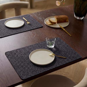 Op de afbeelding: Een eettafel met donkergrijze vilten placemats, crèmekleurige borden en goudkleurige vorken. Een glas water en een bord met brood staan ook op de tafel. De tafel is van donker hout.