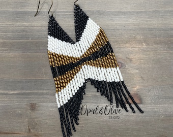 May include: Black, white, and gold beaded fringe earrings with a geometric pattern. The earrings are handmade and feature a unique design. "Opal & Olive Designs" is written on the bottom of the earrings.