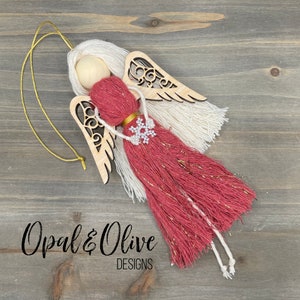 May include: A red and white angel ornament with a wooden cutout of wings and a snowflake charm. The angel has a wooden head and white yarn hair. The ornament has a gold string for hanging. "Opal & Olive Designs" is written on the wooden surface below the ornament.