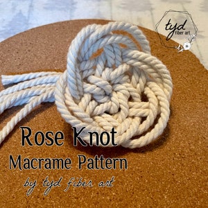 Könnte beinhalten: Eine Nahaufnahme eines weißen Makramee-Rosenknotens auf einer Korkoberfläche. Der Knoten ist aus dickem weißem Seil gefertigt und in einem kreisförmigen Muster gebunden. Der Text "Rose Knot Macrame Pattern by tyd fiber art" ist unter dem Knoten geschrieben.