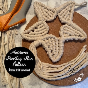 Può includere: Un motivo a stella cadente in macramè beige su una tavola di sughero. La stella è fatta di corda annodata e ha un aspetto strutturato. L'immagine include le forbici e il testo "Macrame Shooting Star Pattern" e "Instant PDF download".