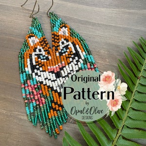 Könnte beinhalten: Ein Paar Perlenohrringe mit einem Tigergesichtsdesign. Die Ohrringe sind aus bunten Perlen in den Farben Orange, Weiß, Schwarz, Türkis und Grün gefertigt. Die Ohrringe sind lang und baumelnd, mit einer Perlenfransen am unteren Ende. Die Ohrringe sind mit "Original Pattern by Opal & Olive Designs" gekennzeichnet.