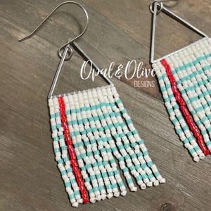 May include: Close-up of a pair of beaded earrings. The earrings feature a silver triangle top with white, turquoise, and red beads. The fringe is made of white beads with turquoise stripes and a red vertical stripe. The earrings are handmade.
