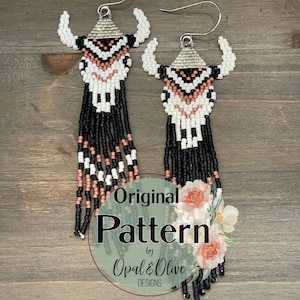 Op de afbeelding: Handgemaakte kralen oorbellen met een stierschedel ontwerp. De oorbellen hebben een zilverkleurige bovenkant, witte hoorns en een patroon van zwarte, witte en roze kralen. Lange zwarte franjes hangen eronder. De tekst "Original Pattern by Opal & Olive Designs" is ook zichtbaar.
