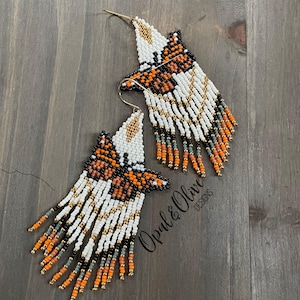 May include: A pair of handmade beaded earrings in a butterfly design. The earrings feature white, orange, black, and gold beads. The design includes a butterfly shape with fringe detailing. The earrings are on a wooden surface.