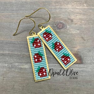 May include: A pair of gold earrings with a rectangular design featuring a pattern of red and green strawberries on a light blue background. The earrings are labeled "Opal & Olive Designs".