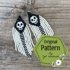 May include: A pair of black and white beaded earrings with a skull design. The earrings are hanging from silver hooks. The earrings are on a wooden surface with a green hexagon sticker that says "Original Pattern by Tyd handmade".