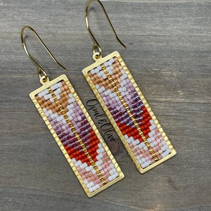 May include: A pair of gold-toned earrings with a rectangular beaded design. The beads are in shades of pink, red, white, and gold. The earrings are made by Opal & Olive.