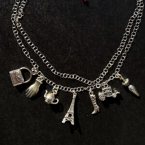 May include: Silver-toned necklace with two strands and multiple charms. Charms include a purse, hand, teapot, Eiffel Tower, boot, carriage, and umbrella. The necklace is displayed on a dark surface.