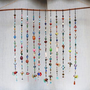May include: A decorative wall hanging featuring multiple strands of colorful beads suspended from a natural wooden branch. The beads are various shapes, sizes, and colors, including red, blue, green, and orange. Some strands have decorative charms at the bottom.