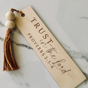 May include: Wooden bookmark with a brown tassel and wooden beads. The bookmark has the text "TRUST in the Lord" and "PROVERBS 3:5-6" engraved on it.