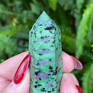 May include: A green, pink and black gemstone point, also known as a crystal, with a smooth, polished surface. The point is held in a hand.