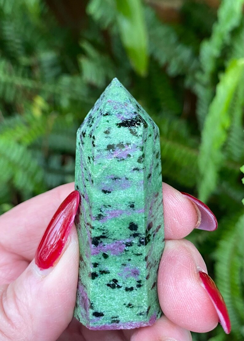 May include: A green, pink, and black gemstone point, also known as a crystal, with a smooth, polished surface. The point is held in a hand.