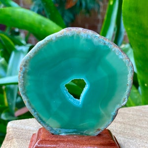 May include: A green agate slice with a natural hole in the center, mounted on a wooden stand. The agate has a smooth, polished surface and a translucent appearance.