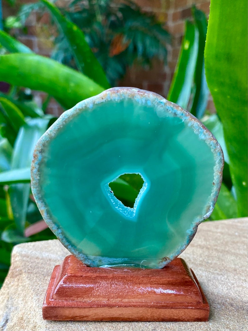 May include: A green agate slice with a natural hole in the center, mounted on a wooden stand. The agate has a smooth, polished surface and a translucent appearance.