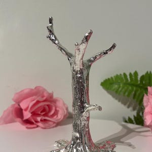 May include: A silver decorative tree sculpture with a textured bark design. The tree has multiple branches extending upwards and a detailed base with leaf-like features. The sculpture is displayed with pink roses and green foliage in the background.