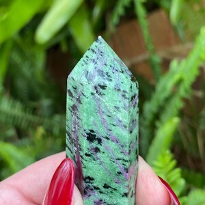 May include: A green, black, and pink zoisite crystal point. The crystal is smooth and polished, with a pointed tip. It is a natural stone and has a unique pattern of colors.