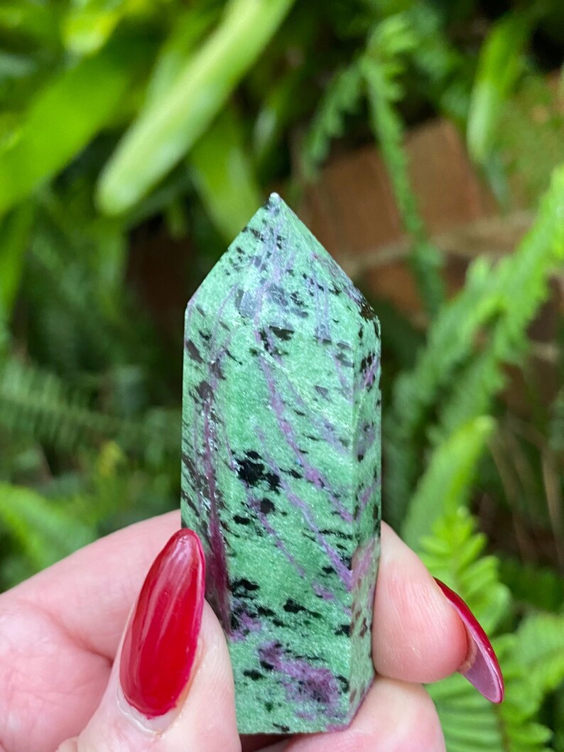 May include: A green, black, and pink zoisite crystal point. The crystal is smooth and polished, with a pointed tip. It is a natural stone and has a unique pattern of colors.