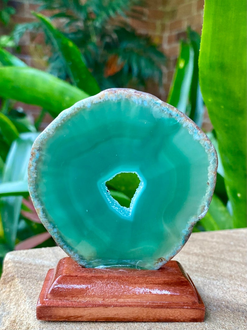May include: A green agate slice with a hole in the center, displayed on a wooden stand. The agate has a natural, polished finish.