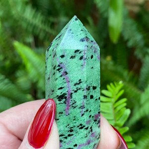 May include: A green, black, and purple gemstone point, also known as a crystal, with a faceted shape. The point is held in a hand with red nail polish.