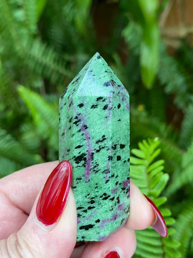 May include: A green, black, and purple gemstone point, also known as a crystal, with a faceted shape. The point is held in a hand with red nail polish.