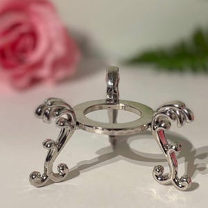 May include: A silver-coloured decorative stand with an open circular top and ornate, curved legs. The stand has a small, raised support and is designed to hold a round object. The background includes a blurred pink flower and green foliage.