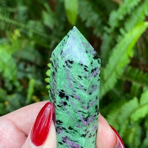 May include: A green, black, and purple gemstone point, also known as a crystal, is held in a hand. The point is smooth and polished.