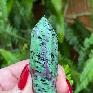 May include: A green, black, and pink zultanite crystal point. The crystal is faceted and has a pointed tip. It is held in a hand with red nail polish.