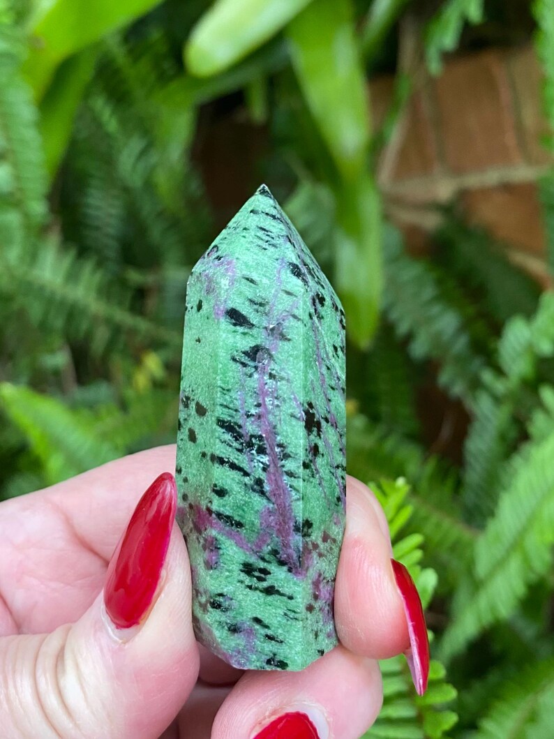 May include: A green, black, and pink zultanite crystal point. The crystal is faceted and has a pointed tip. It is held in a hand with red nail polish.