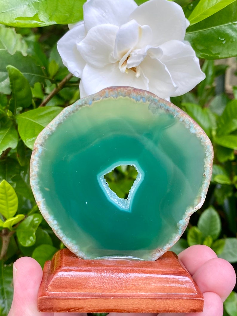 May include: A green agate slice with a natural hole in the center, displayed on a wooden stand.