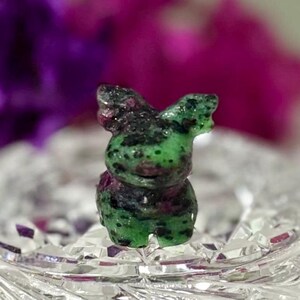 May include: A small, carved gemstone figurine of a bunny. The bunny is mainly green with black and red inclusions. It rests on a clear, faceted glass surface, with a blurred purple and pink background.