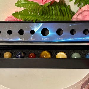 May include: A black box set containing gemstone planets. The planets are arranged in a row, with the names Mercury, Venus, Earth, Mars, Jupiter, Saturn, Uranus, and Neptune printed above. The gemstones vary in colour.