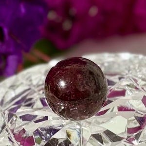 May include: A deep red, spherical bead or decorative item sits on a clear, cut-glass surface. The background shows blurred purple flowers, providing a contrast. The overall effect is one of elegance and detail.