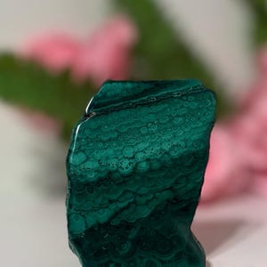 May include: A polished malachite stone with a rich, deep green color and intricate swirling patterns. The natural stone has a smooth, glossy surface and a unique, organic shape. The malachite is displayed against a soft, blurred background.