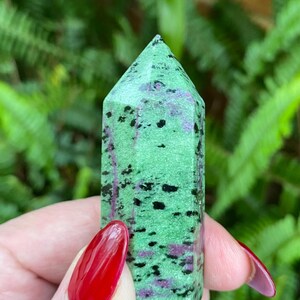 May include: A green, black, and purple gemstone point with a pointed tip. The gemstone is polished and has a smooth surface.