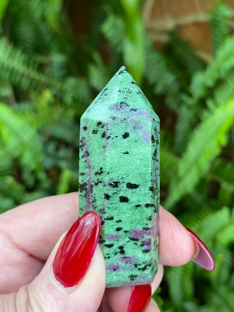 May include: A green, black, and purple gemstone point with a pointed tip. The gemstone is polished and has a smooth surface.