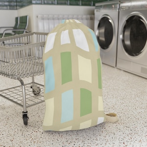 May include: A drawstring laundry bag featuring a beige background with a repeating geometric pattern of vertical rectangles in white, light blue, pale yellow, and sage green. The bag has a cinched top with a drawstring closure at the opening.