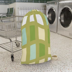 May include: A drawstring laundry bag featuring a repeating geometric pattern of vertical rectangles in shades of olive green, white, light cream, and pale aqua blue. The bag has a cinched top closure with a beige cord and is standing upright.