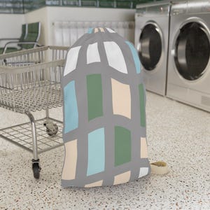 May include: A drawstring laundry bag featuring a gray grid pattern with alternating rectangular blocks of white, light blue, sage green, and beige. The bag has a cinched top closure and is constructed from fabric with a repeating geometric design.