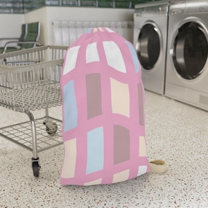 May include: A cylindrical laundry bag featuring a pink base color with a repeating pattern of vertical rectangular blocks in shades of white, light blue, beige, and muted brown. The bag has a drawstring closure at the top and a fabric strap.