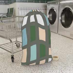 May include: A drawstring laundry bag featuring a grid pattern of dark gray lines over rectangular blocks of white, light blue, forest green, and beige. The bag has a cylindrical shape with a cinched top and a small beige cord visible at the base.