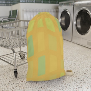 May include: A drawstring laundry bag featuring a repeating grid pattern of vertical and horizontal rectangles in shades of mustard yellow, light yellow, and pale lime green. The bag has a cinched top closure with a beige fabric cord.