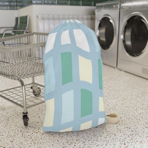 May include: A laundry bag featuring a light blue grid pattern with alternating rectangular blocks in white, pale yellow, and seafoam green. The bag has a drawstring closure at the top and is made of fabric, designed for holding clothing items.