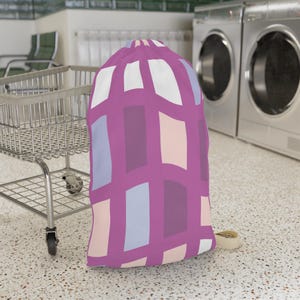 May include: A drawstring laundry bag featuring a grid pattern of magenta lines filled with rectangular blocks in shades of white, light blue, pale pink, and dark purple. The bag has a cinched top closure and a beige fabric drawstring cord at the base.