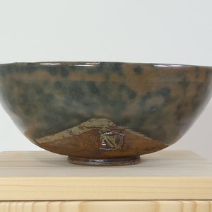 Può includere: Una ciotola in ceramica marrone con una glassa verde. La ciotola ha un bordo largo e un fondo arrotondato. La ciotola è appoggiata su una superficie di legno.